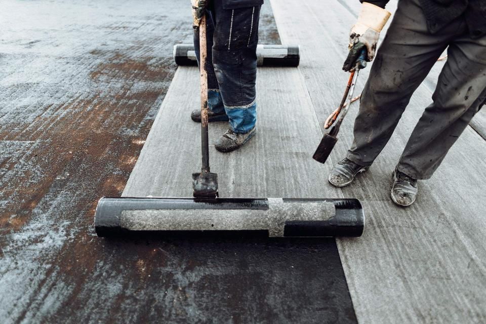 Two workers installing roofing material, one using a roller, the other with a torch.