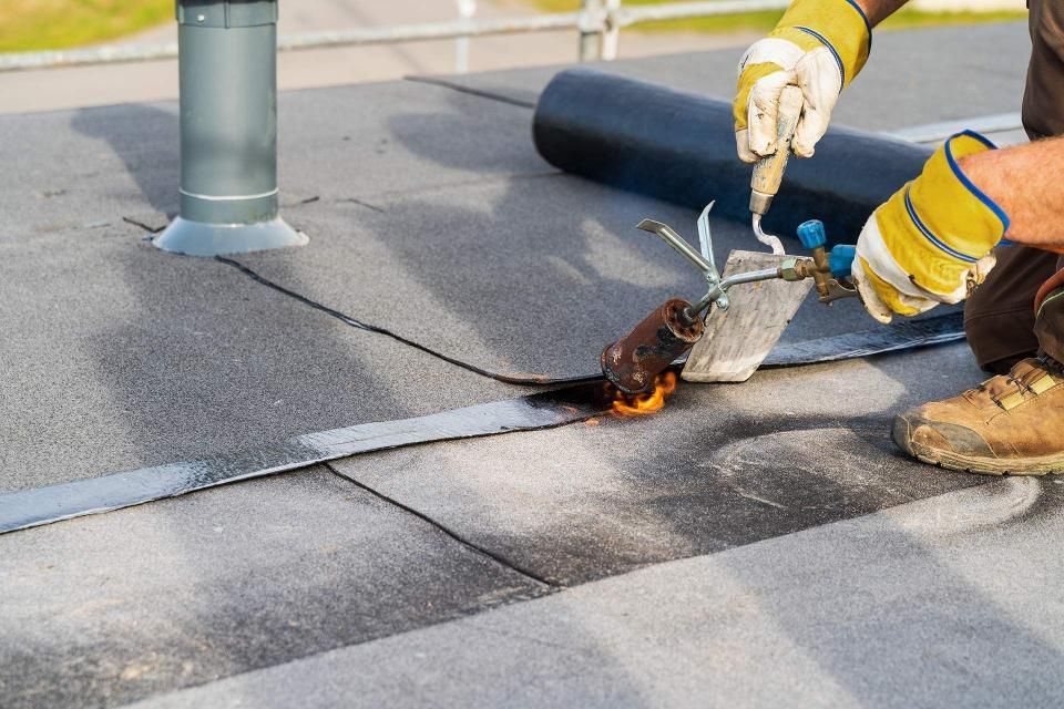 Roofer using a torch to seal seams on a flat, gray roof; hands in yellow gloves, brown work boots, black rolled roofing nearby.