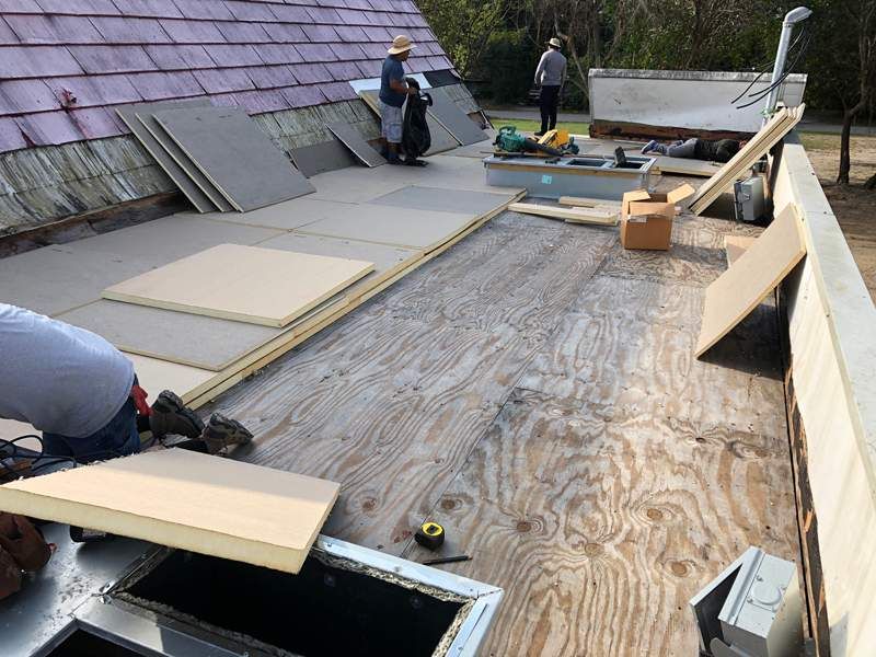 Workers installing roof insulation and plywood on a building's flat roof; tools and materials are scattered around.