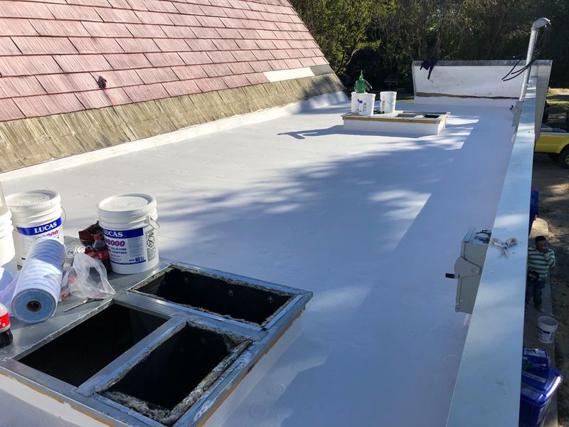 White roof being worked on, with materials visible.  A wooden building with a brown shingle roof is adjacent. Sunny day.