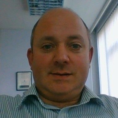 A man with a shaved head wearing a light blue and white striped shirt, smiling slightly in an office setting.