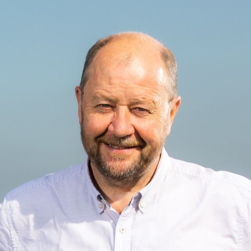 A man with a short beard smiling, wearing a light-colored button-up shirt against a plain, light blue background.