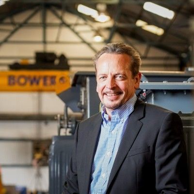 A smiling person in a suit and checkered shirt stands in a large industrial factory with yellow machinery in the back.