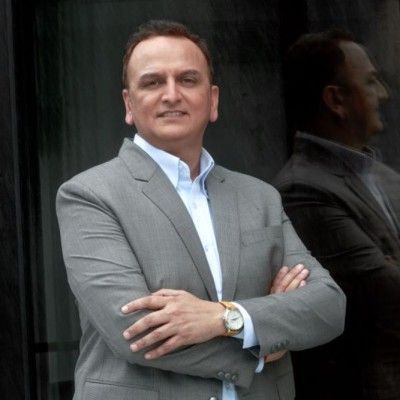 A man in a light grey blazer and white collared shirt stands with arms crossed against a dark, reflective background.