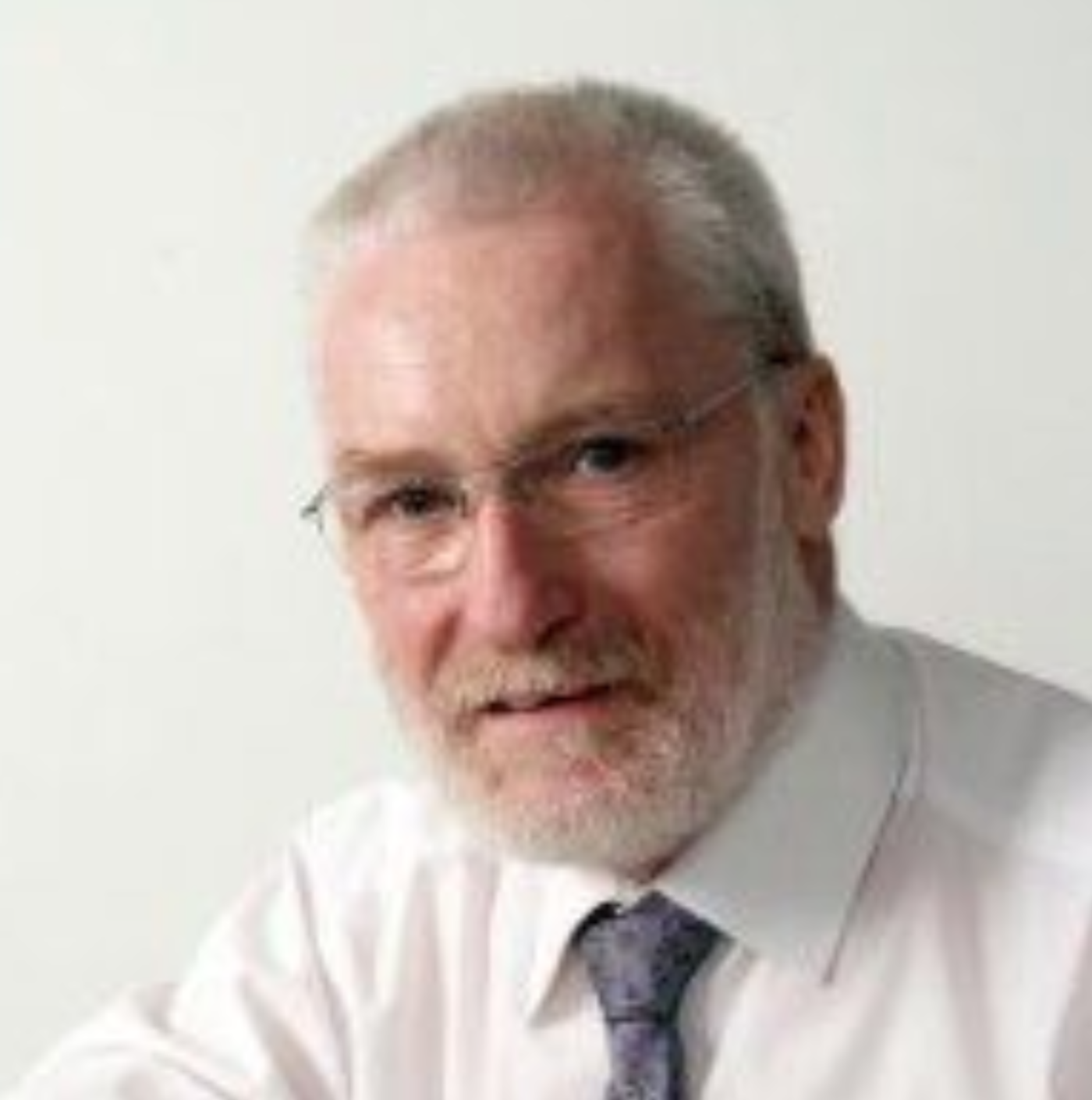 A person with short grey hair and a beard, wearing glasses, a white collared shirt, and a grey patterned necktie.
