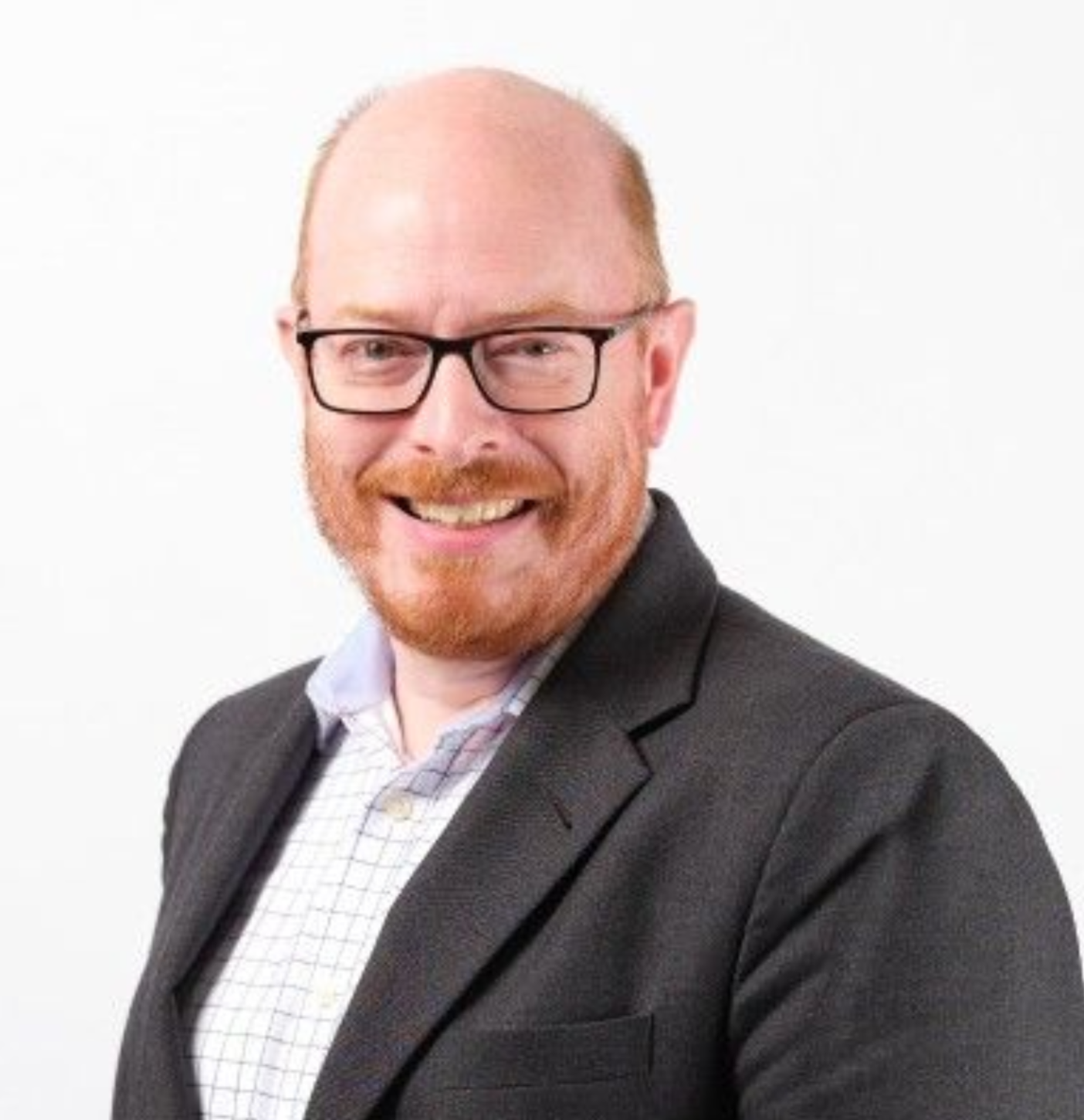 A smiling man with a red beard and glasses, wearing a grey blazer over a checkered dress shirt against a white background.