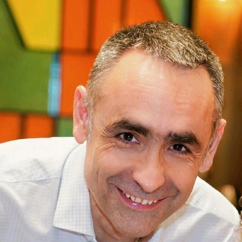 A close-up portrait of a smiling person wearing a white collared shirt against a blurred, colorful background.