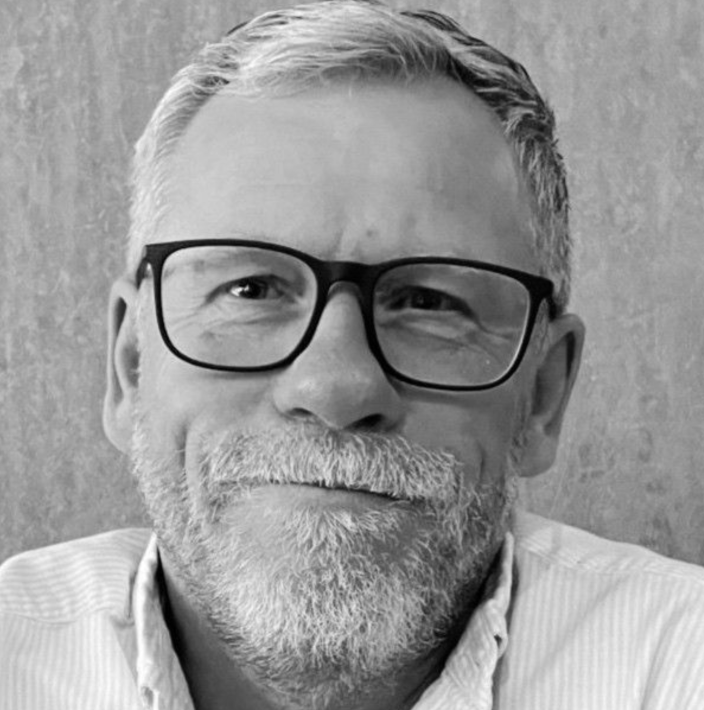 A headshot of a person with a short beard and glasses, wearing a collared shirt, smiling softly against a textured wall.
