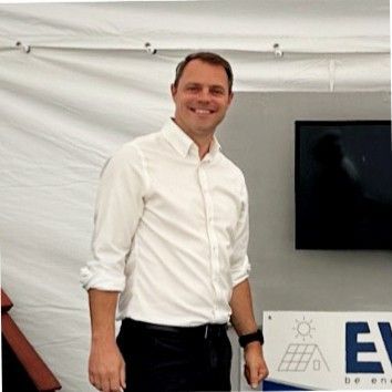 A person smiling in a white shirt and dark pants stands before a sign with a solar panel icon and part of a brand name.