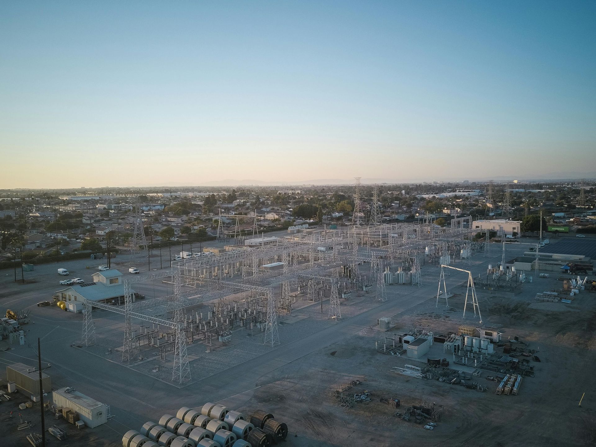 Aerial view of a large electrical substation with metal towers and equipment, set against a sunset sky and suburban area.