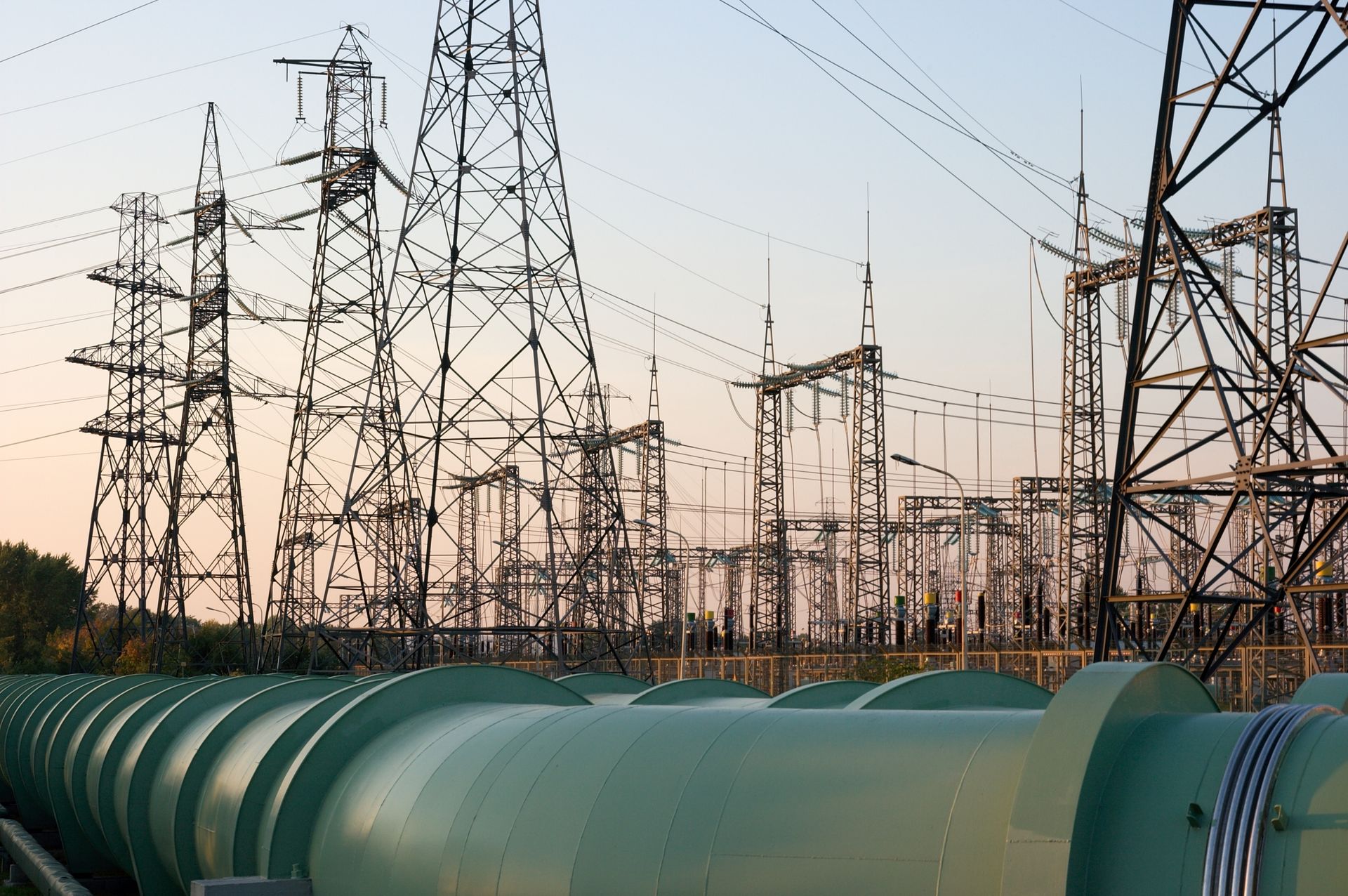 Large green industrial pipes in the foreground against a backdrop of metal electrical transmission towers at sunset.