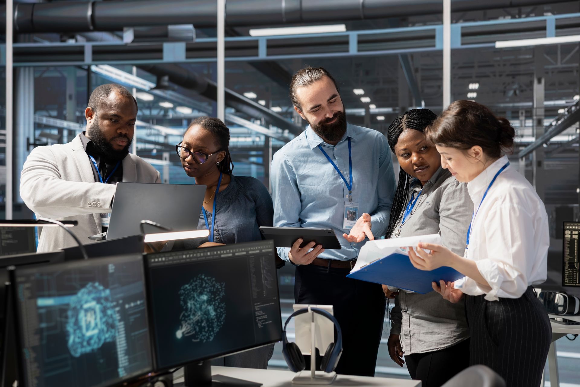 A diverse team of professionals collaborates around computer monitors and a document in a modern, industrial-style office.