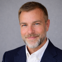 Headshot of a man with short brown hair and a beard, wearing a white collared shirt and blue blazer against a grey backdrop.