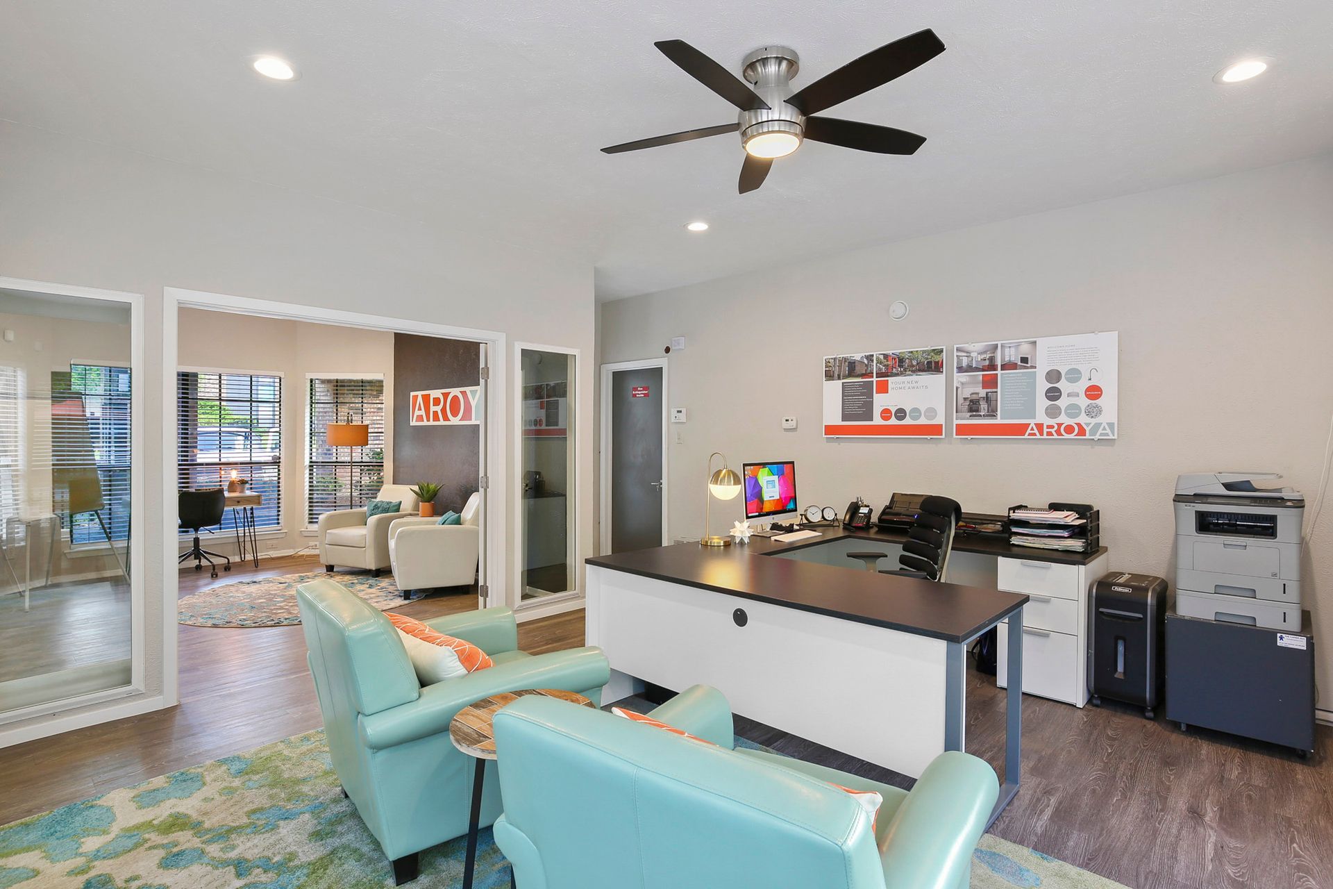 Office interior with desk, chairs, and ceiling fan; a second room visible in the background.