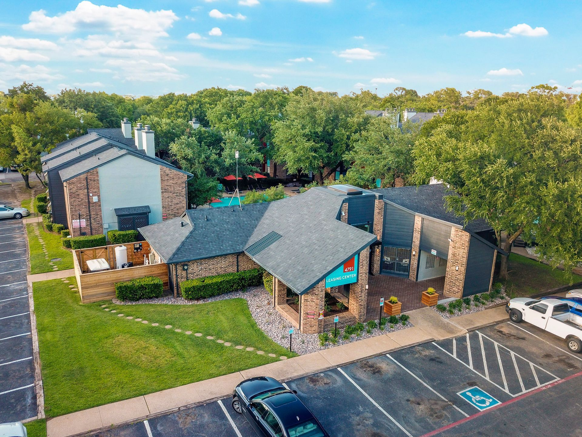 Aerial view of an apartment complex with a pool, parking lot, and green trees.