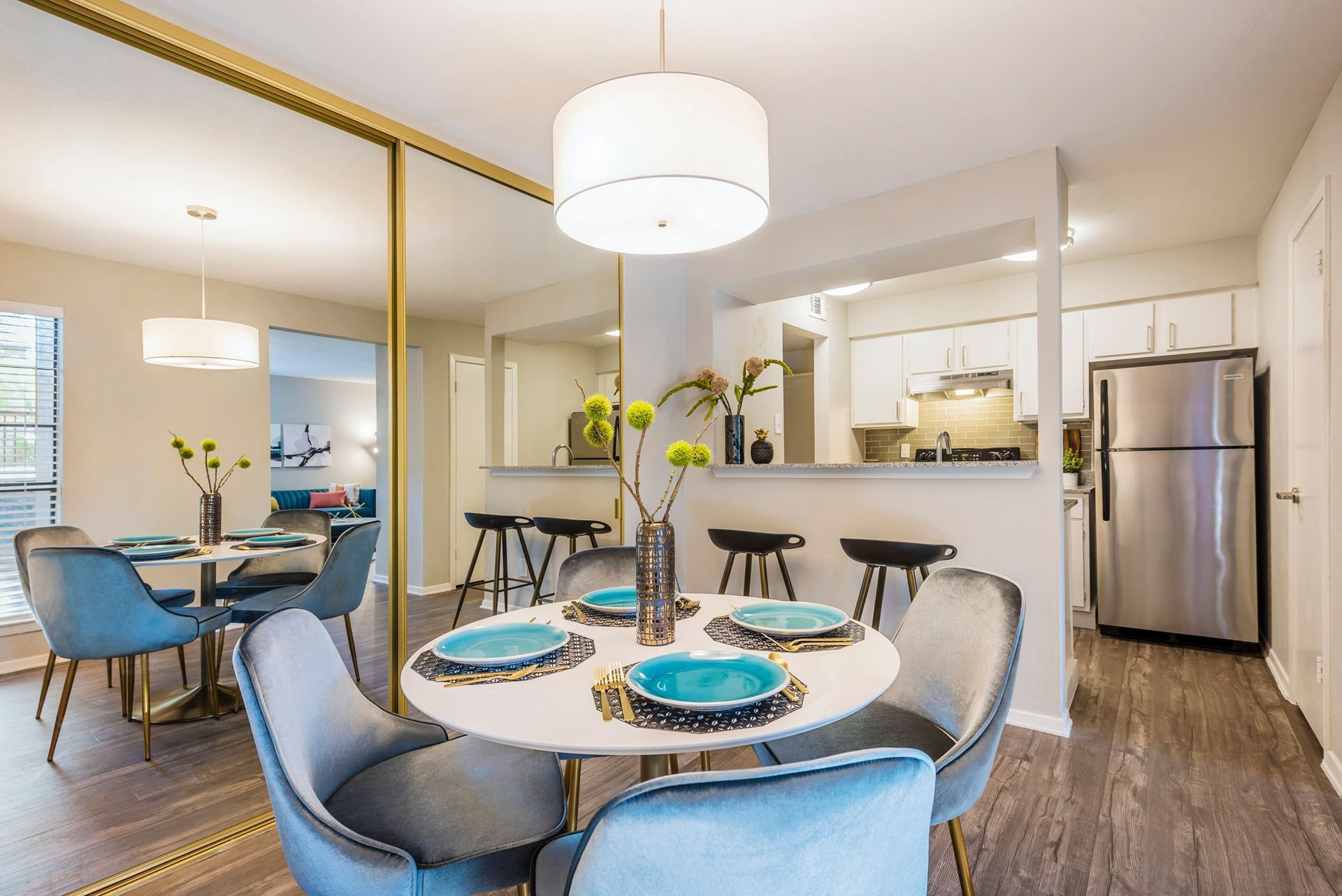 Dining room with white round table, teal chairs, and large mirror. Kitchen visible in the background.
