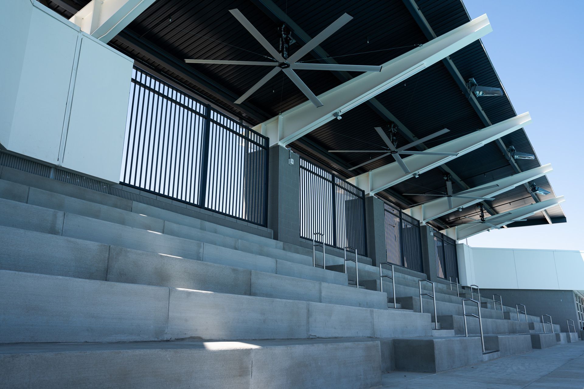 Empty concrete bleachers under a covered area with black corrugated roof and large ceiling fans.