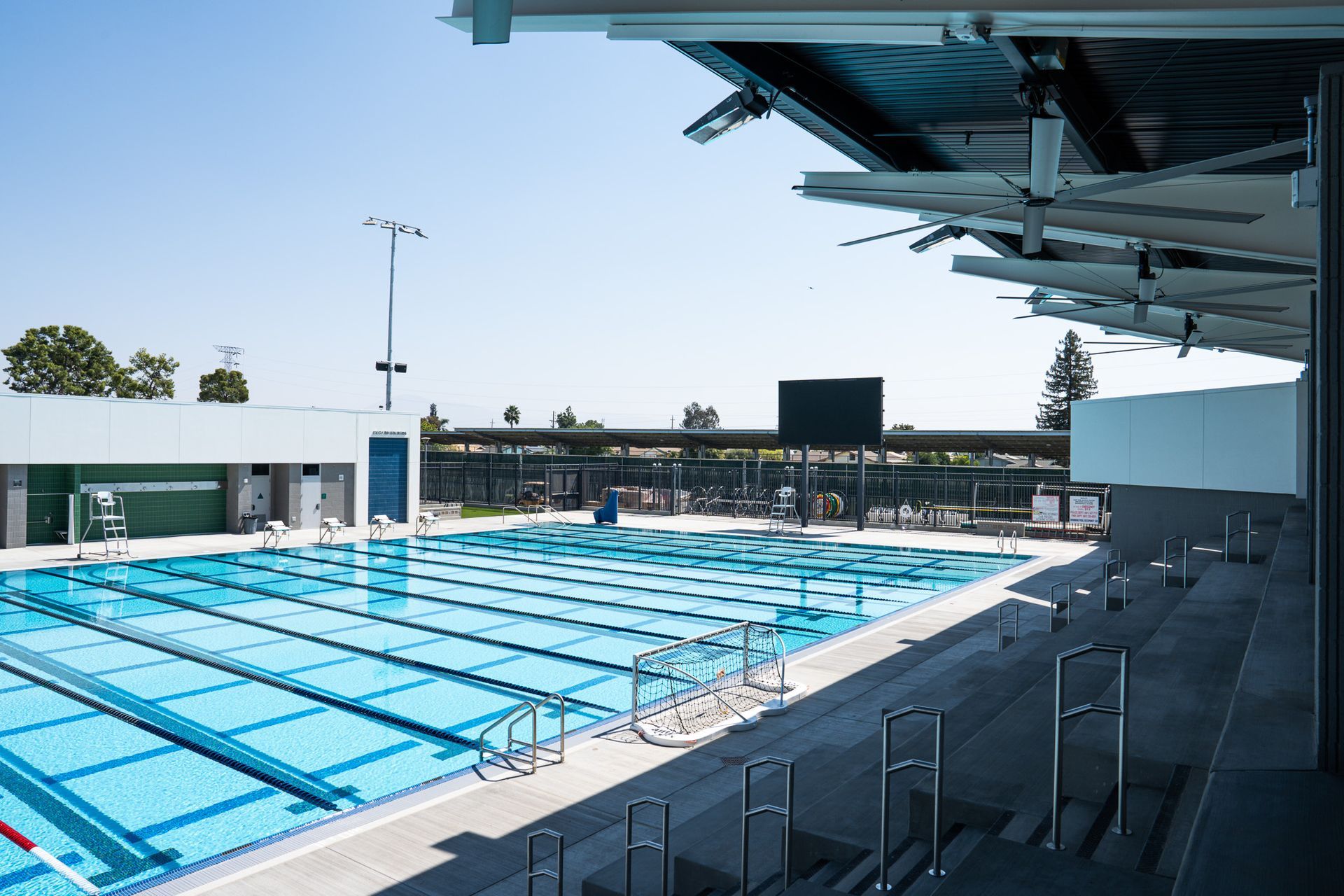 Swimming pool with lanes, bleachers, and overhead shade structure under a clear sky.
