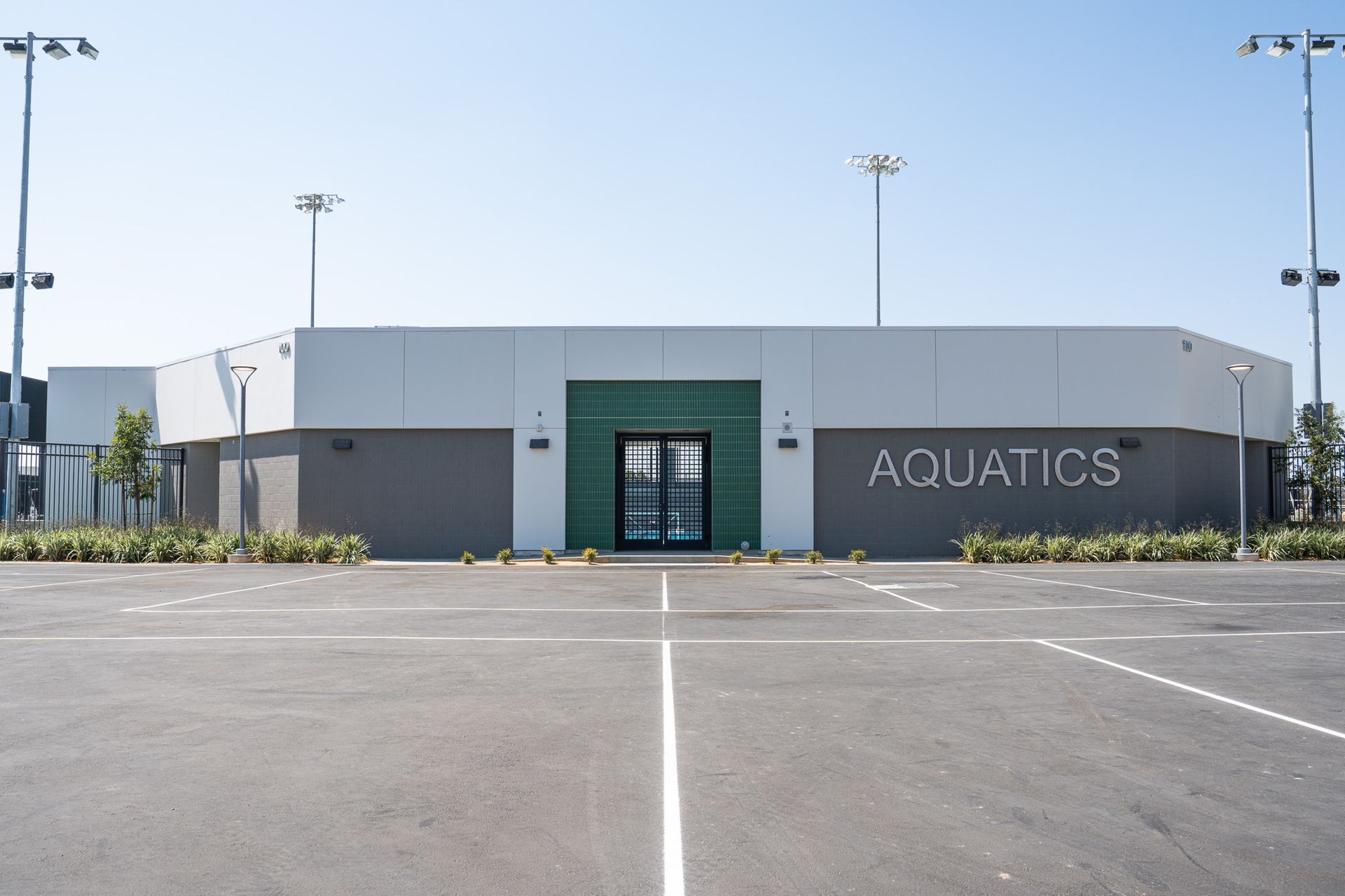 Aquatics center with a parking lot. Light gray and white building, green doors, and the word 