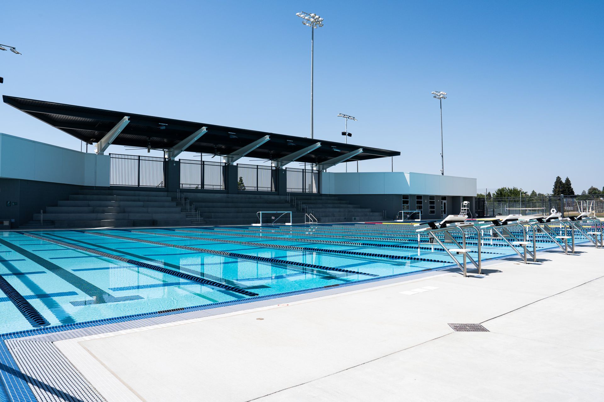 Outdoor swimming pool with blue water, concrete deck, seating, and a covered structure under a bright blue sky.