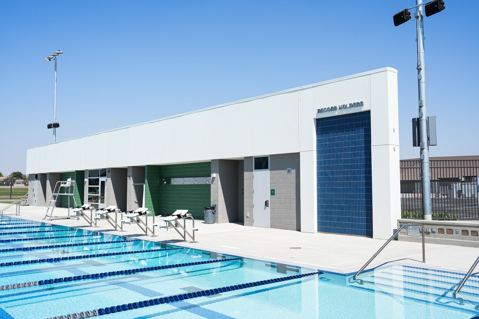 A rectangular swimming pool with lanes, adjacent to a white building with a blue door, under a clear sky.