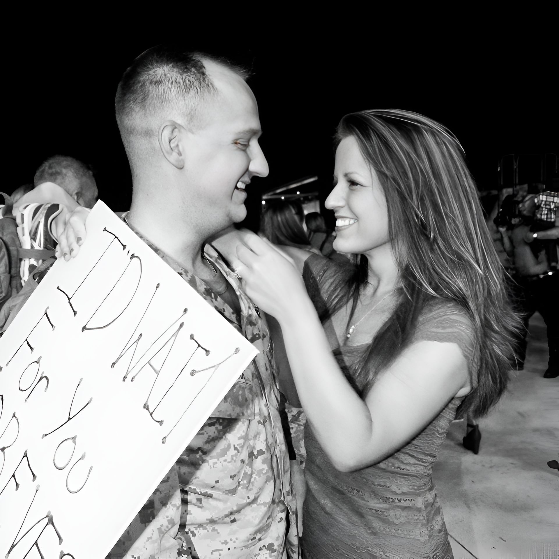 Smiling couple outdoors at night, woman adjusting the man's shirt while he holds a sign.