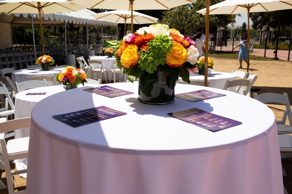 Round tables with white tablecloths, flower centerpieces, and printed materials, set outdoors under umbrellas.