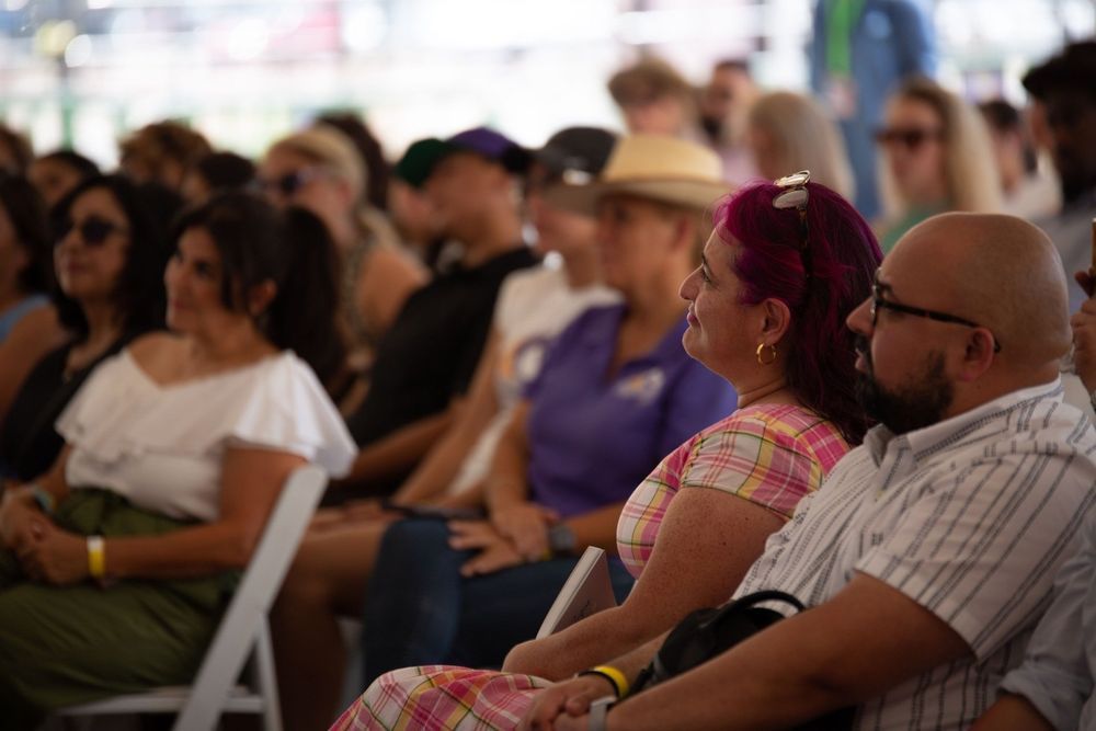 People seated outdoors, watching something. Some wearing sunglasses, one with pink hair.