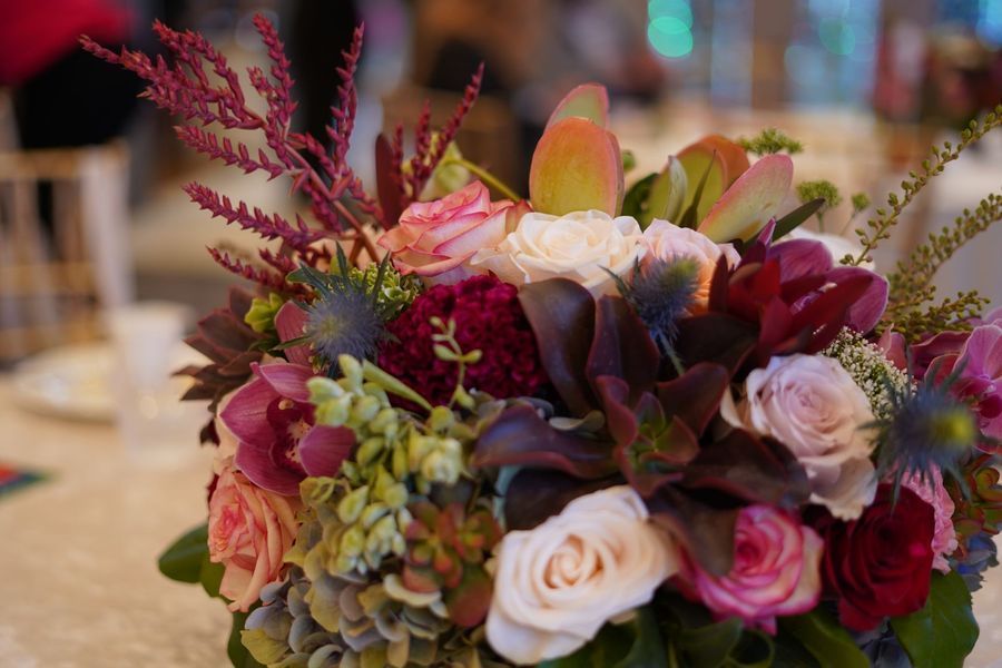 Close-up of a vibrant floral arrangement with pink and purple roses, blue thistles, and greenery on a white tablecloth.