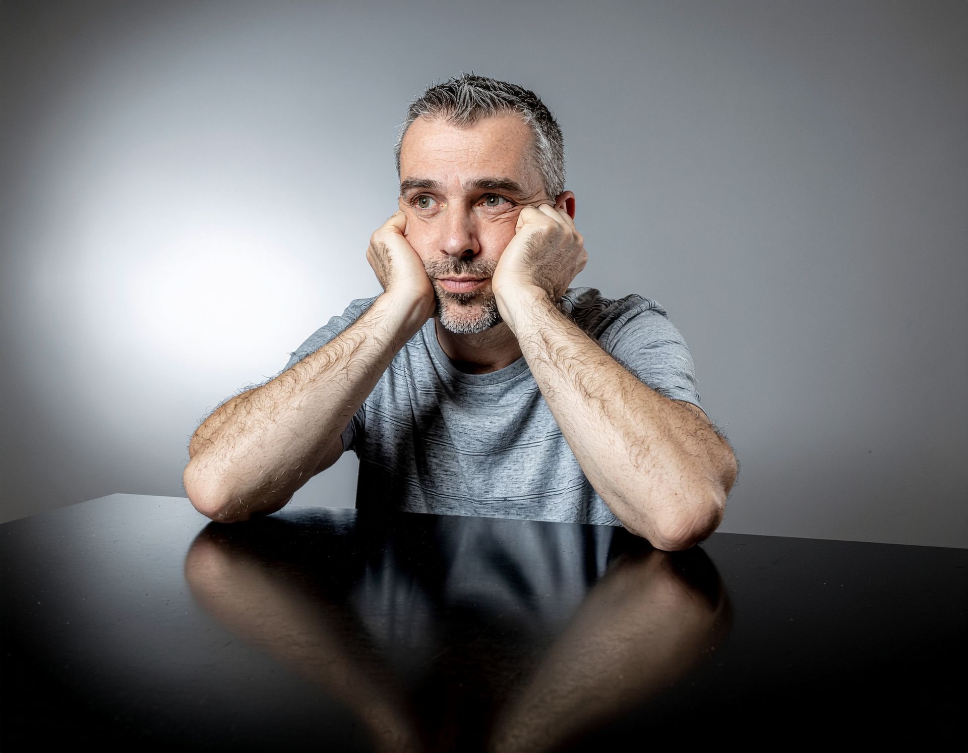 Man resting his chin on hands, looking despondent. Sitting at a dark reflective table. Gray background and t-shirt.