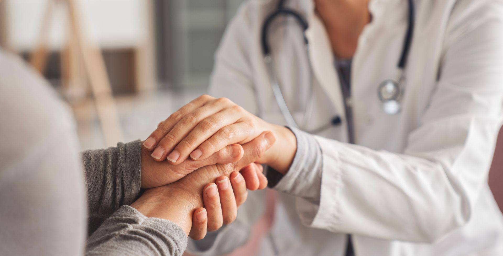 Doctor's hands comforting a patient's hands. Doctor wears a white coat and stethoscope.
