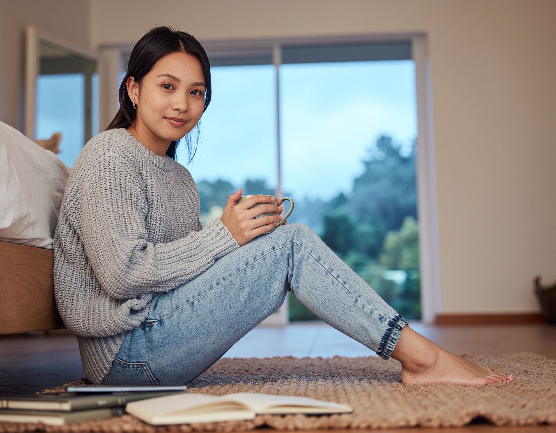 Woman sitting on a rug with a mug, reading near a large window with a book in the foreground