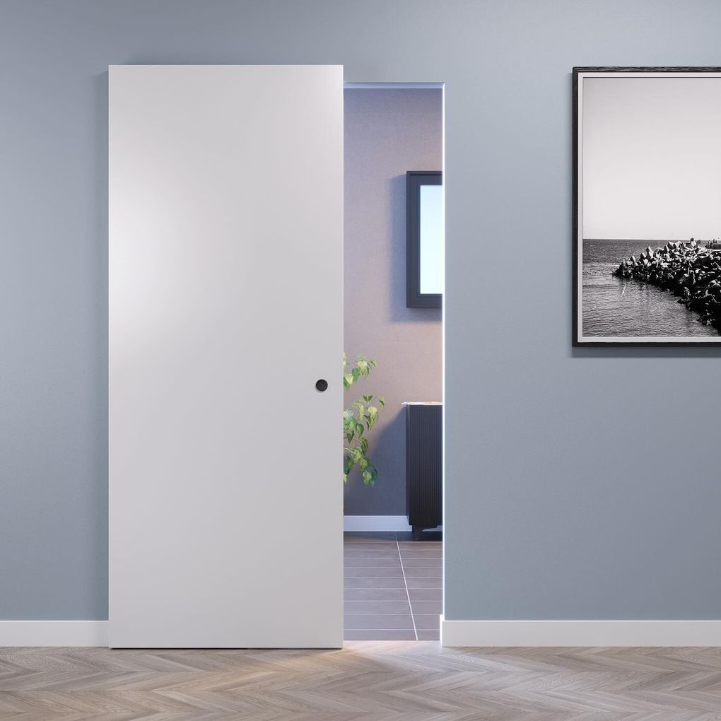 Una porta bianca socchiusa in una stanza grigia lascia intravedere un corridoio piastrellato e una fotografia in bianco e nero incorniciata.