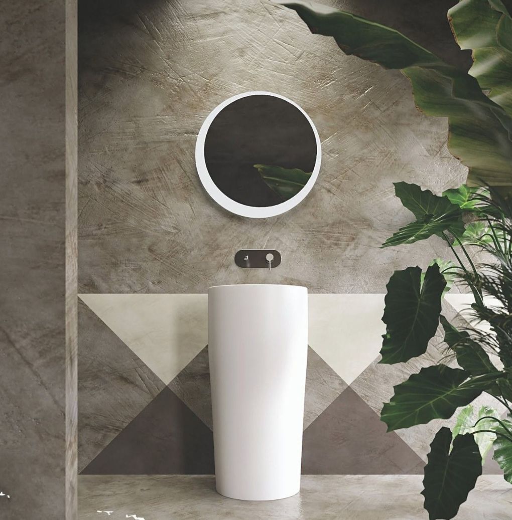 Bagno moderno con specchio rotondo sopra un lavabo a colonna bianco e piastrelle geometriche a parete, con una pianta sulla destra.