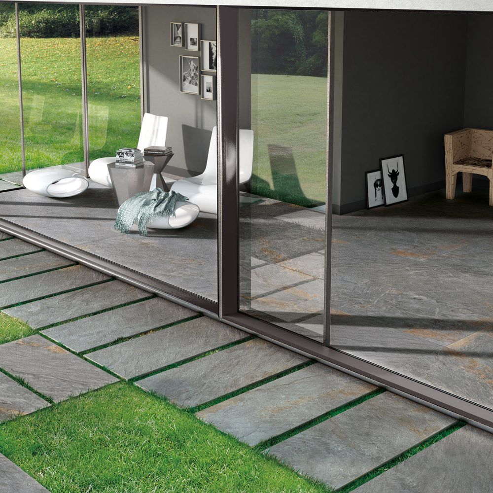 Patio moderno con ampie porte scorrevoli in vetro, pavimentazione in pietra, prato verde e arredamento esterno minimalista.
