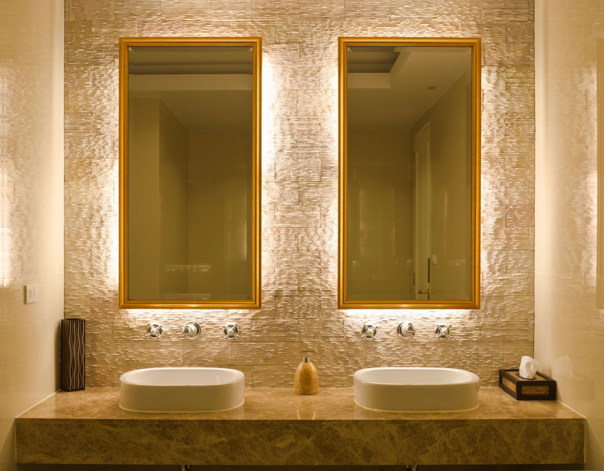 Elegante mobile da bagno con due lavabi rotondi, specchi con cornice dorata e illuminazione calda su una parete con finitura materica.