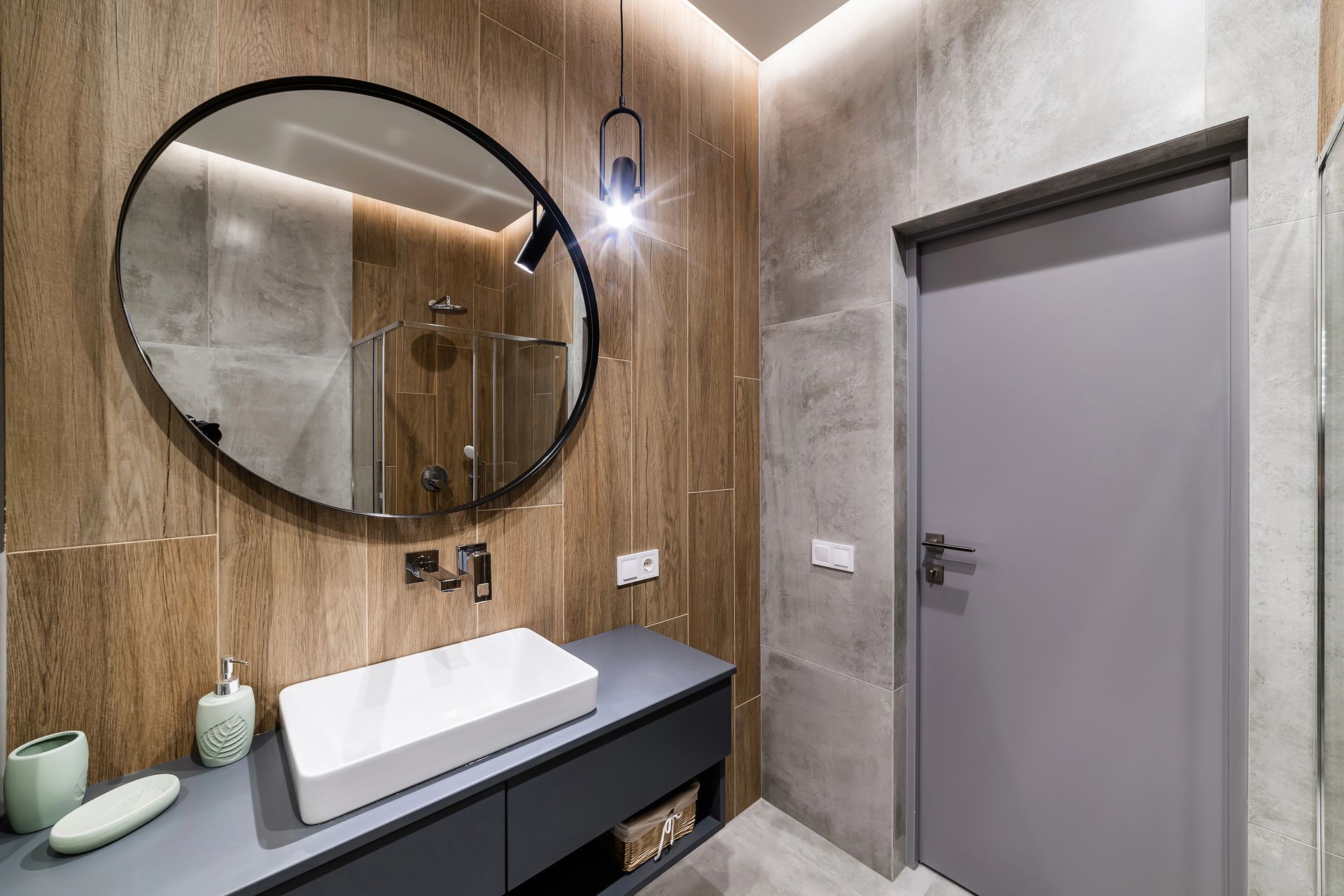 Mobile bagno moderno con specchio rotondo, parete in legno, lampada a sospensione e anta grigia.