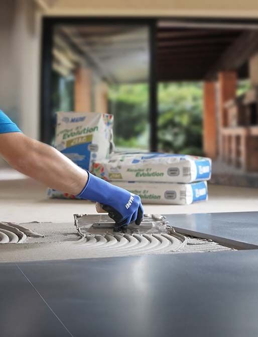 Mano guantata che stende l'adesivo sul pavimento con una cazzuola accanto a sacchi da costruzione impilati.