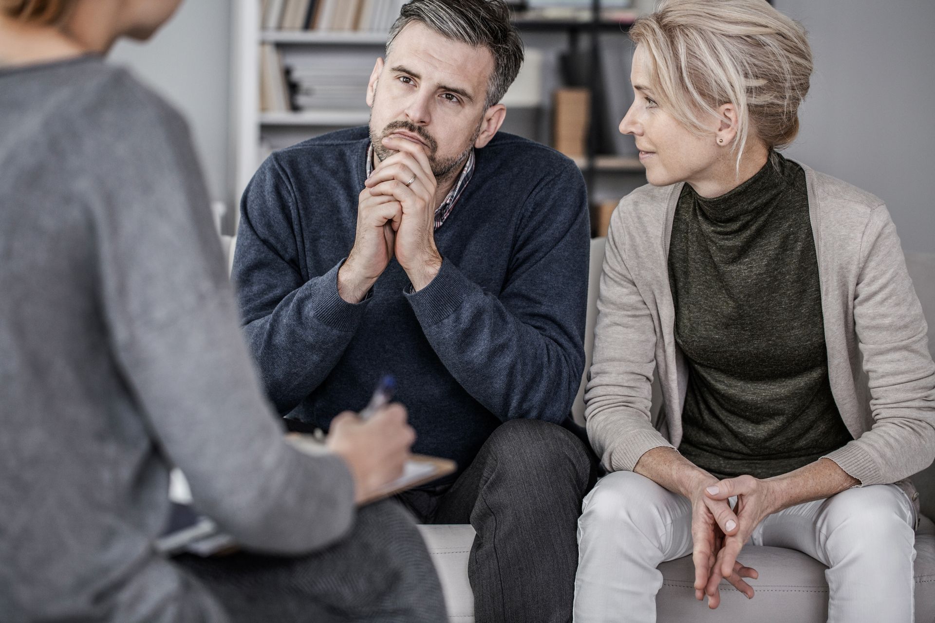 A couple sitting together on a couch, talking thoughtfully, with a counselor taking notes nearby