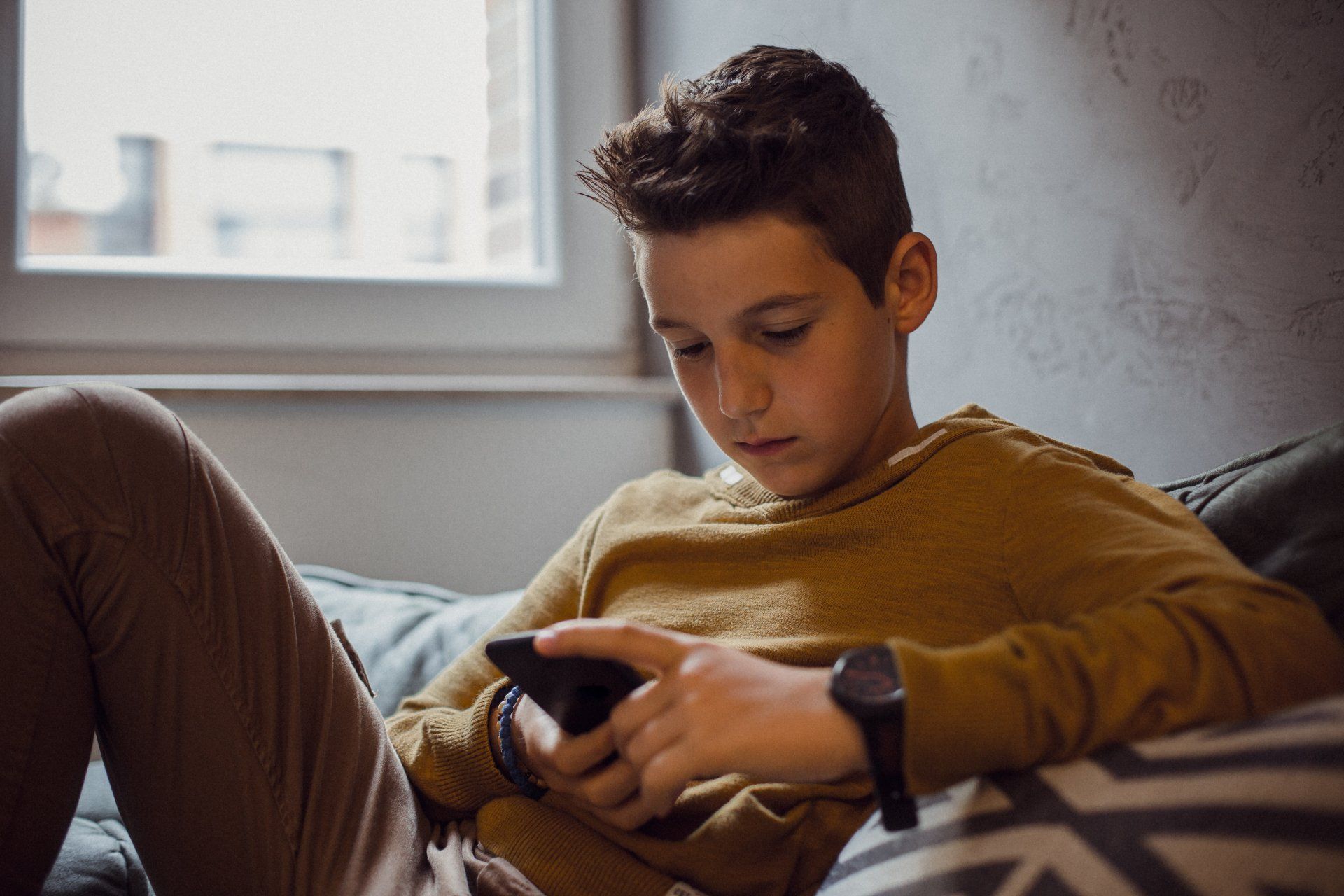Boy on a couch looking at a phone. He is wearing a yellow sweater and smartwatch.