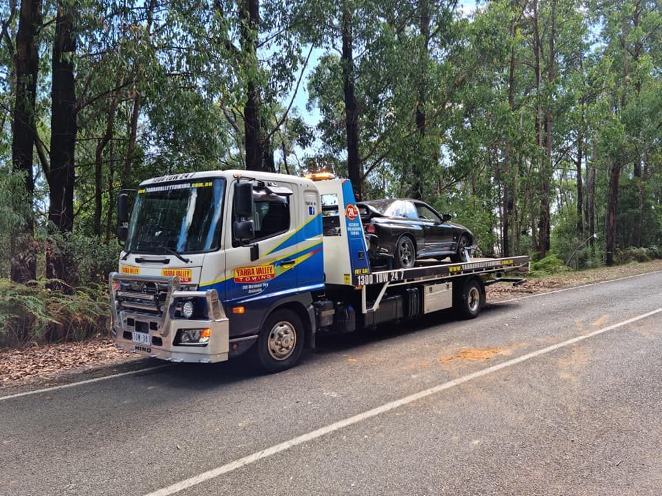 Tow truck with a black car on its bed, parked on a road lined with trees.