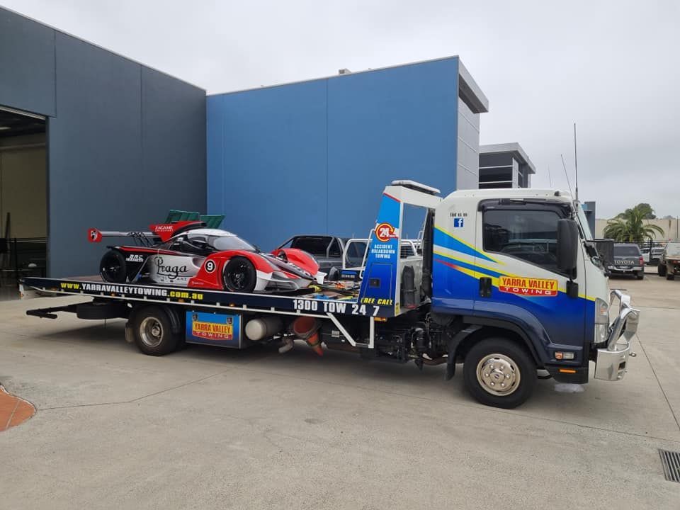 A tow truck carrying a red and white race car outside a building on a cloudy day.