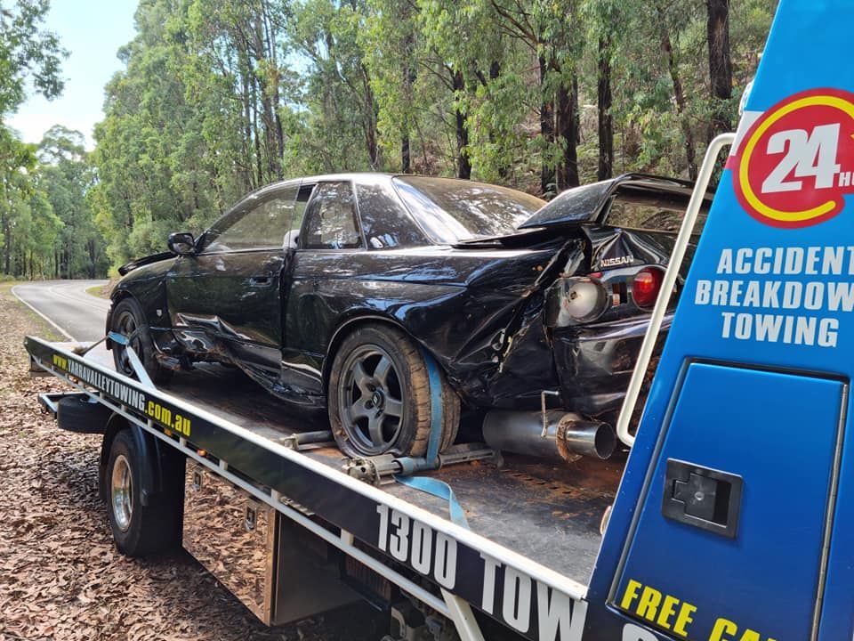 Black car badly damaged on a tow truck, roadside setting, with forest in the background.