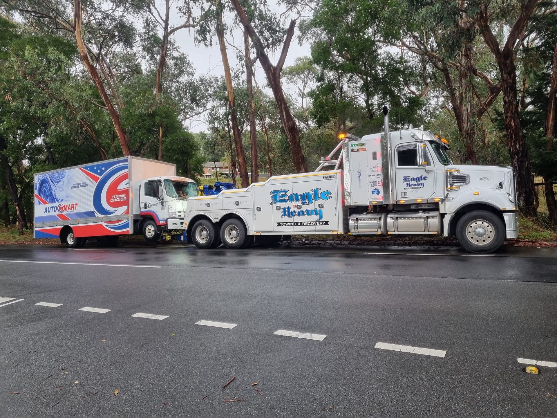 A tow truck hauls a box truck on a tree-lined roadside. The trucks are white with blue lettering.