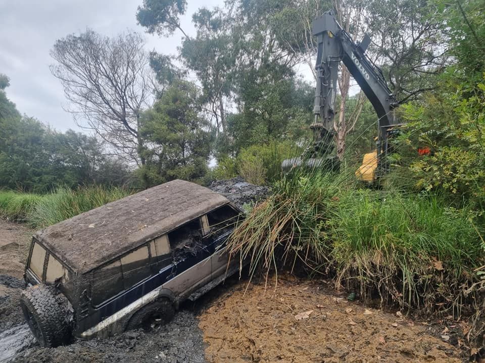 Muddy SUV stuck in a ditch being pulled out by an excavator. Gray vehicle, brown mud, green foliage.