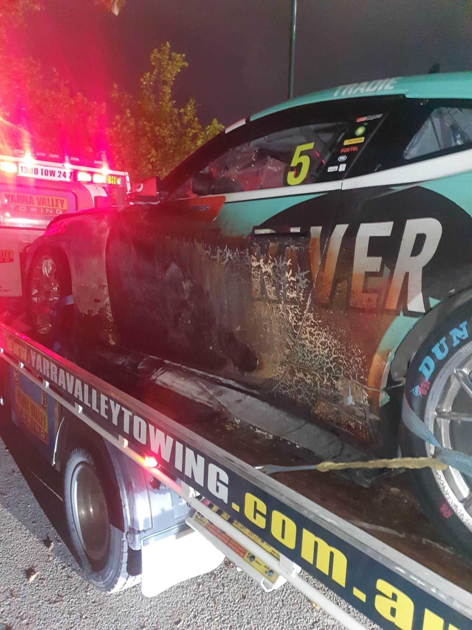 Race car on a tow truck; covered in debris; flashing red lights.