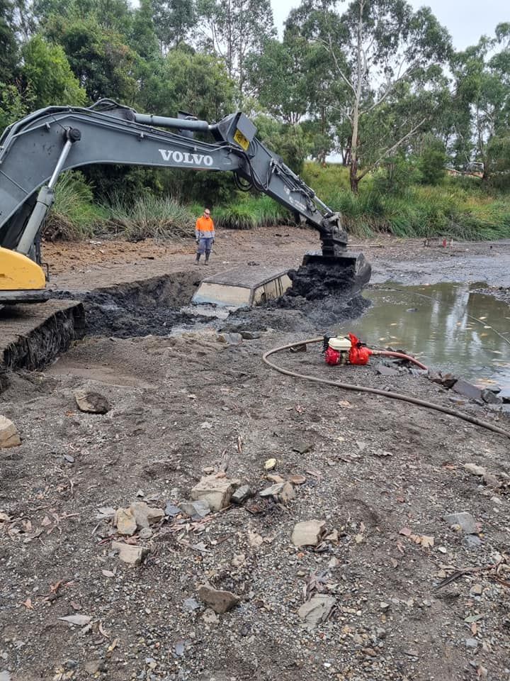 An excavator scoops dark material from a muddy area. A worker stands nearby.
