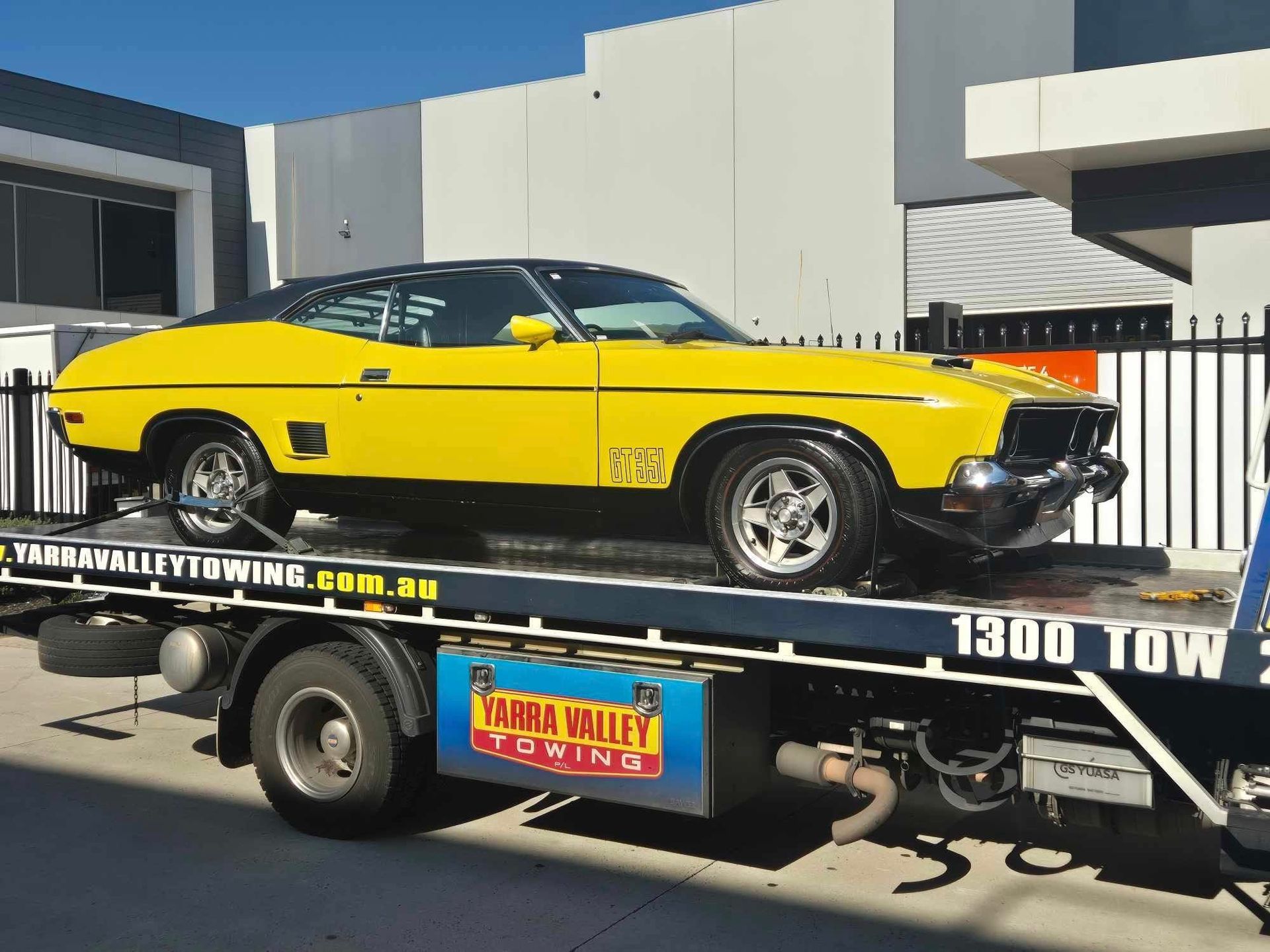 Yellow classic car on a tow truck, next to a building. 