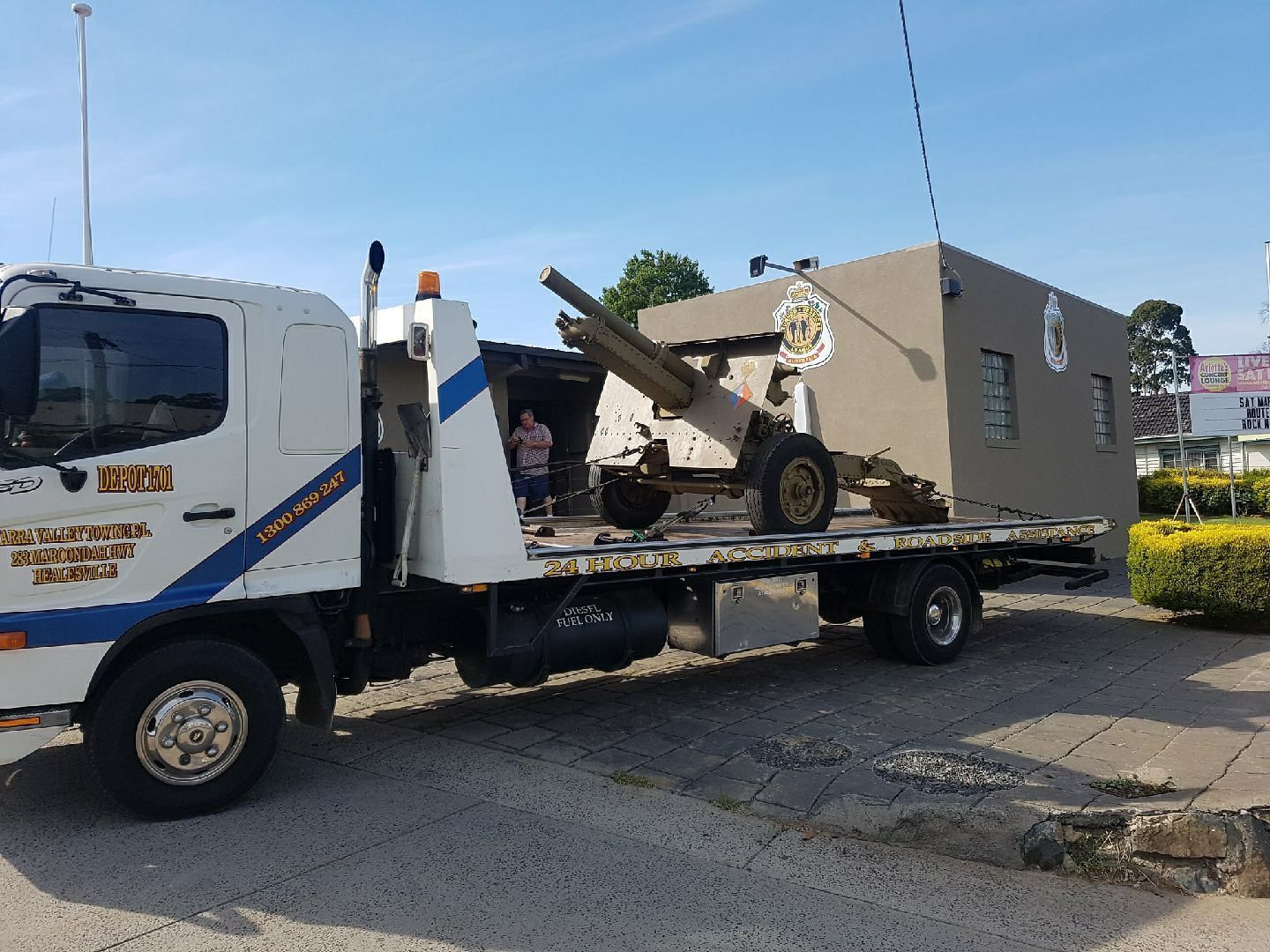 A flatbed tow truck transporting a military cannon and a small building outdoors on a sunny day.