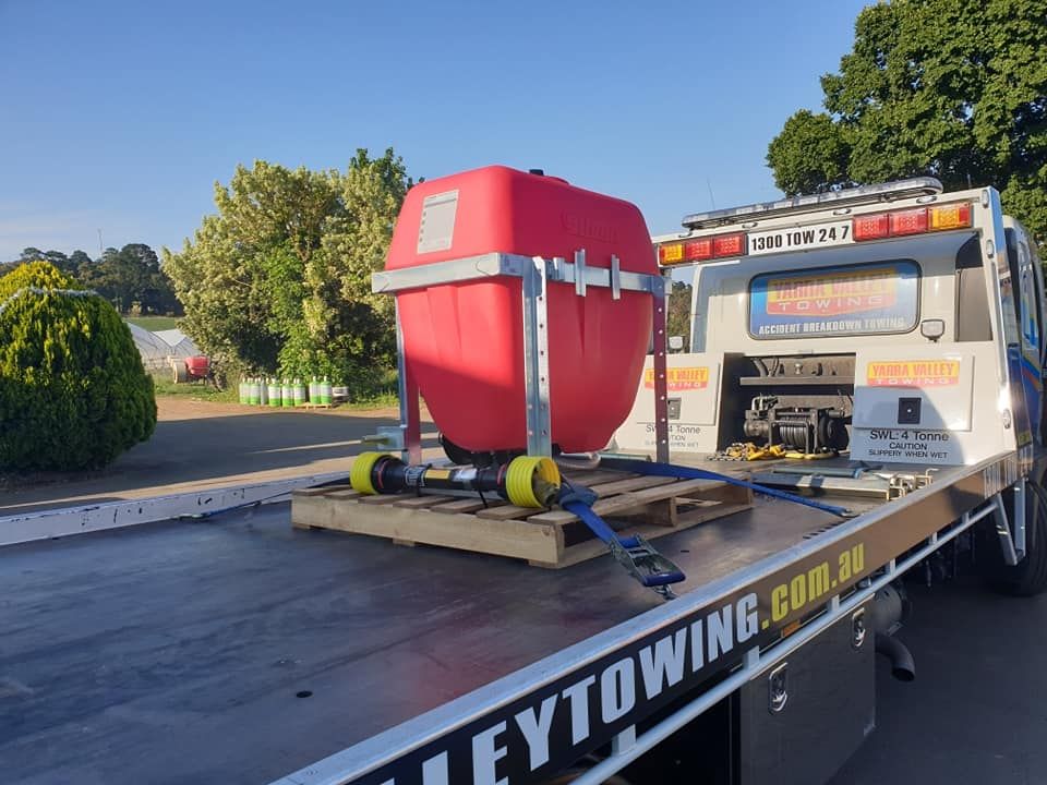 Red tank secured on a flatbed tow truck. Sunny outdoor setting with trees in the background.